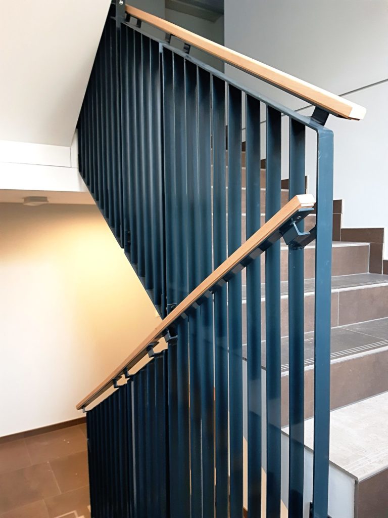 Vnitřní antracitové schodišťové zábradlí se svislou výplní z ploché ocele s kolmou orientací. Zábradlí je osazené dubovým madlem.