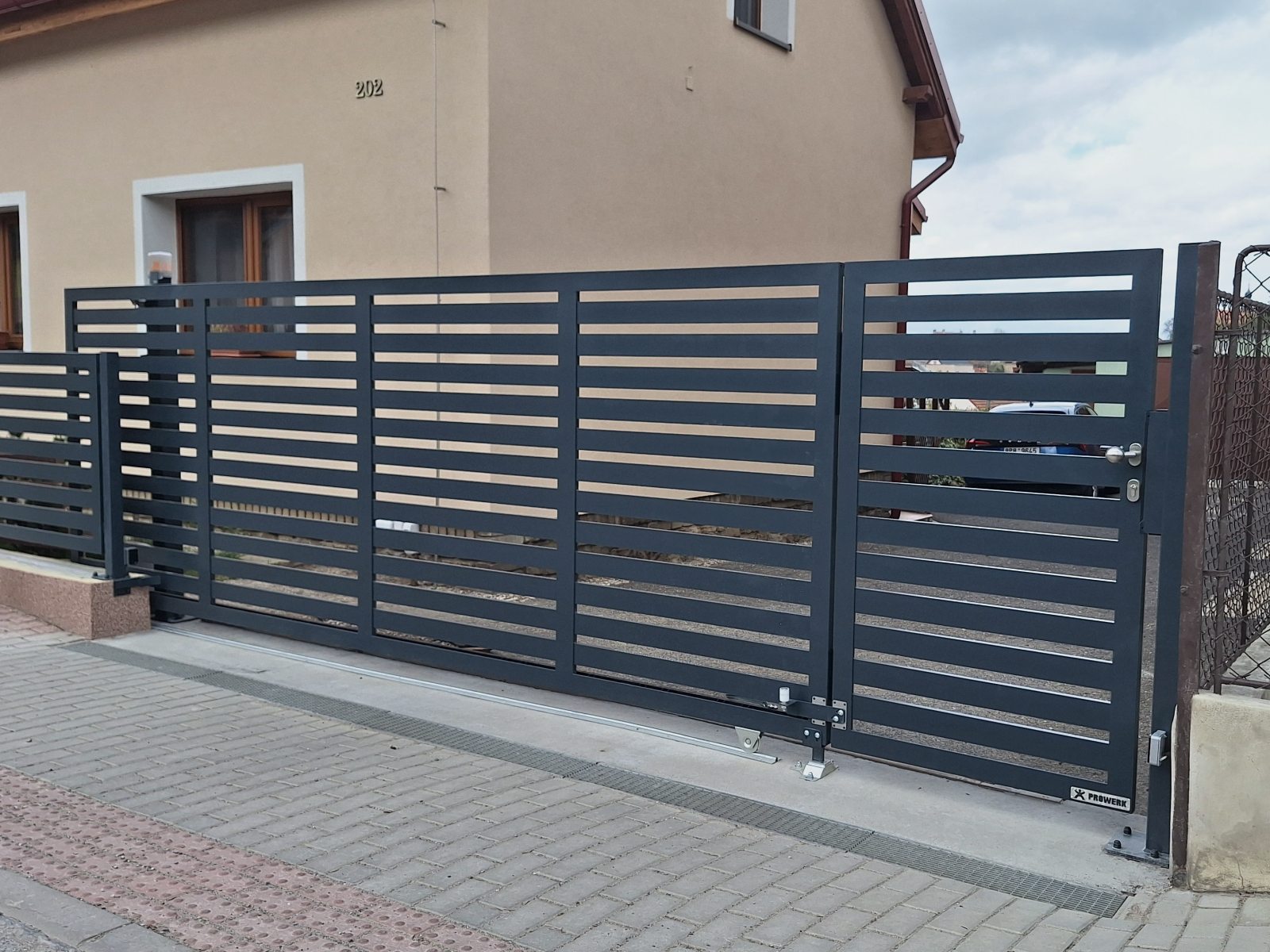 Pojezdová brána na kolejnici PROWERK s integrovanou brankou SYNERGY. Výplň brány vodorovné hliníkové lamely šířky 80mm. Lakováno v odstínu antracit. Realizace okres Beroun.